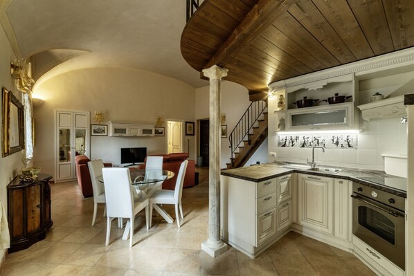 Una cocina amplia con muebles blancos, un comedor con mesa de vidrio y una escalera que sube a un nivel superior.