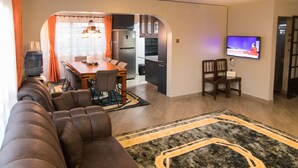 Un salon doté d’un grand canapé d’angle, un coin repas avec une table et des chaises, une cuisine équipée d’un réfrigérateur et un téléviseur fixé au mur.