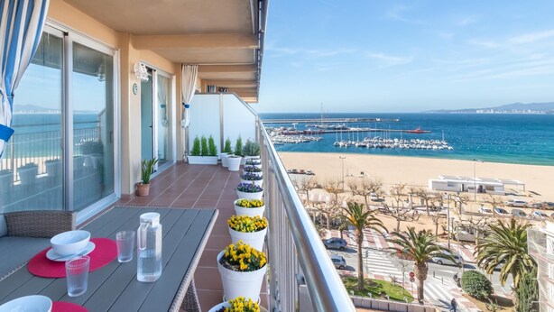 Un balcon avec vue sur la mer, des plantes en pot et une table à manger dressée avec des assiettes et des verres.