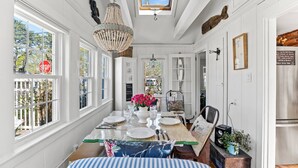 A dining area with a table set for four, a chandelier, and a view of the outdoors through large windows. - Hosting and Helping Other making Memories managed by Itrip Property Management (Barnstable)