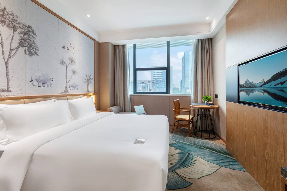 Et hotelværelse med en stor seng, et skrivebord med en bærbar computer, et fjernsyn og udsigt over byen.
