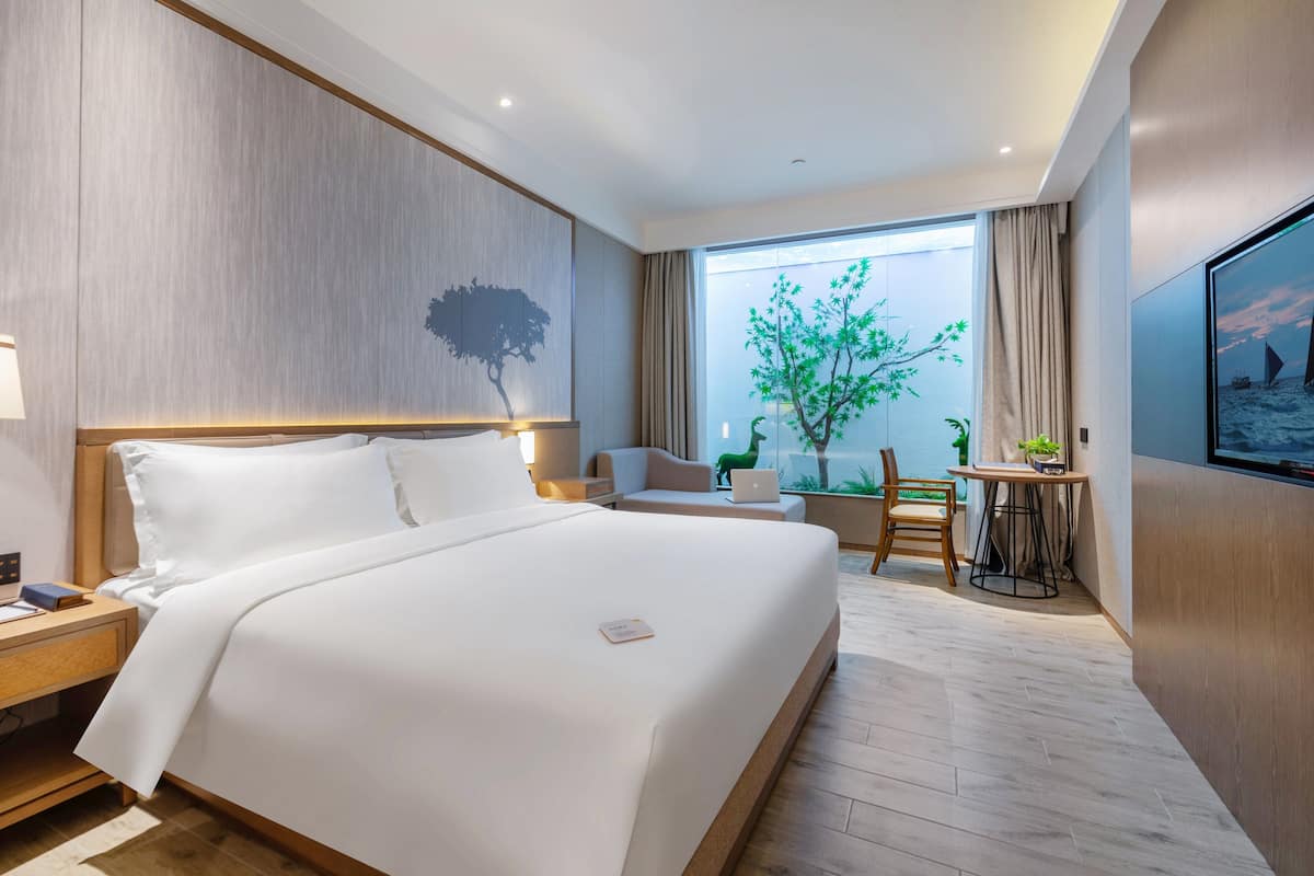 Et moderne hotelværelse med en stor seng, et skrivebord med en bærbar computer, en stol og udsigt til et træ gennem et vindue.