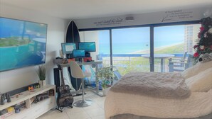 Una camera da letto con un letto, una scrivania con due monitor, una chitarra e vista sulla spiaggia.