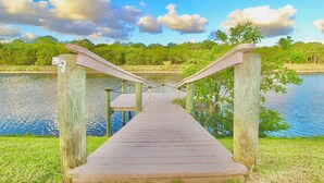 Un puente de madera sobre un lago tranquilo, con cielo despejado y vegetación al fondo.