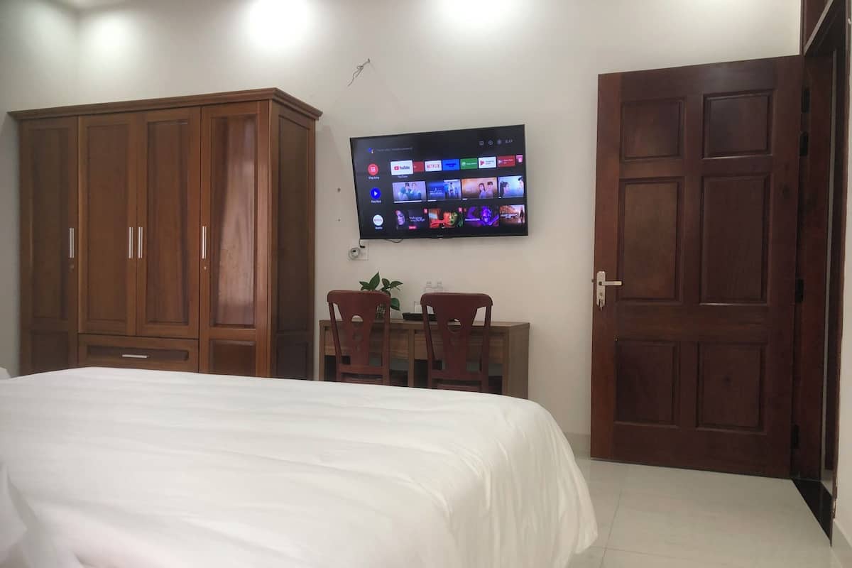 A bedroom with a bed, wooden wardrobe, a TV mounted on the wall, and a desk with chairs.