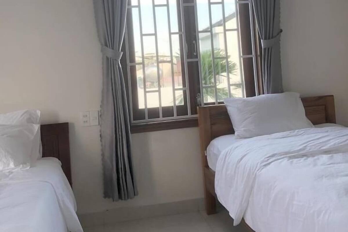 A hotel room with two single beds, a large window with vertical blinds, and a view of the outside.
