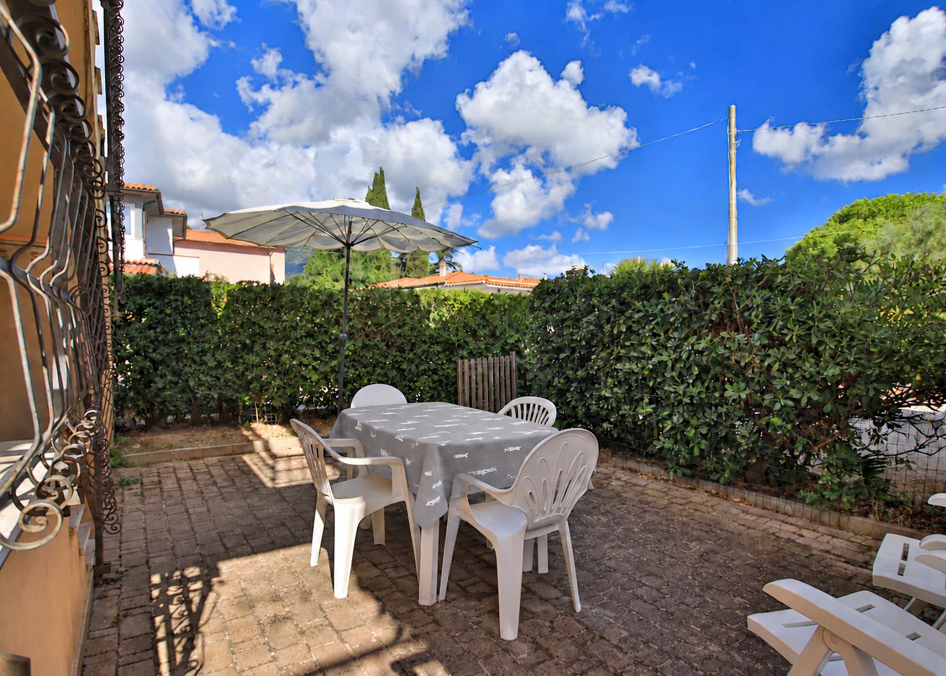 An outdoor patio with a table and chairs, a pergola with a white umbrella, and a hedge in the background.
