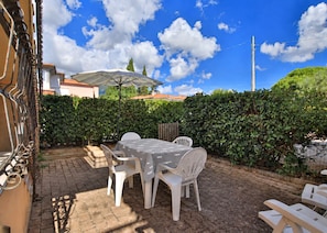 Une terrasse extérieure avec une table et des chaises, une pergola surmontée d’un parasol blanc et une haie en arrière-plan.