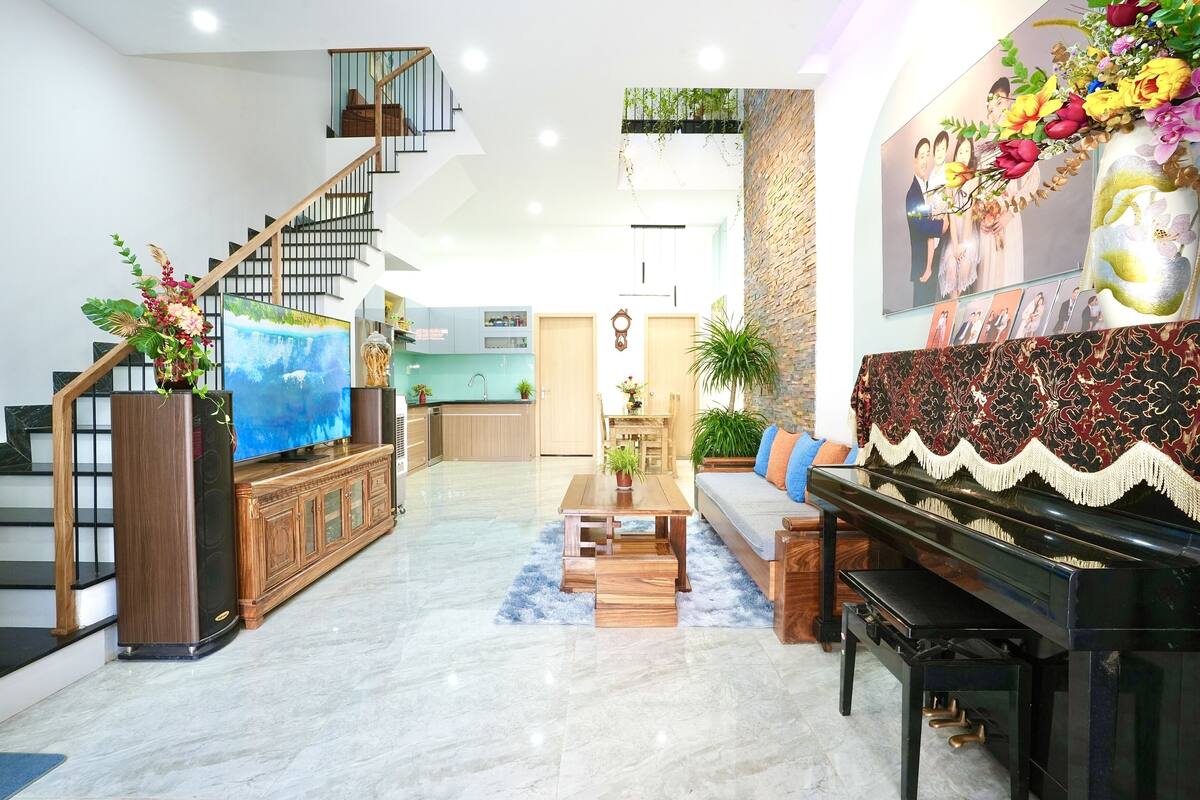 A spacious living area with a staircase, a piano, a television, and a kitchen in the background.