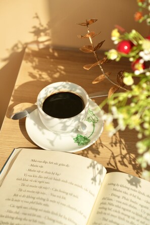 A cup of coffee on a saucer with a spoon, next to an open book on a wooden table. - KALINH HOUSE (Da Nang)