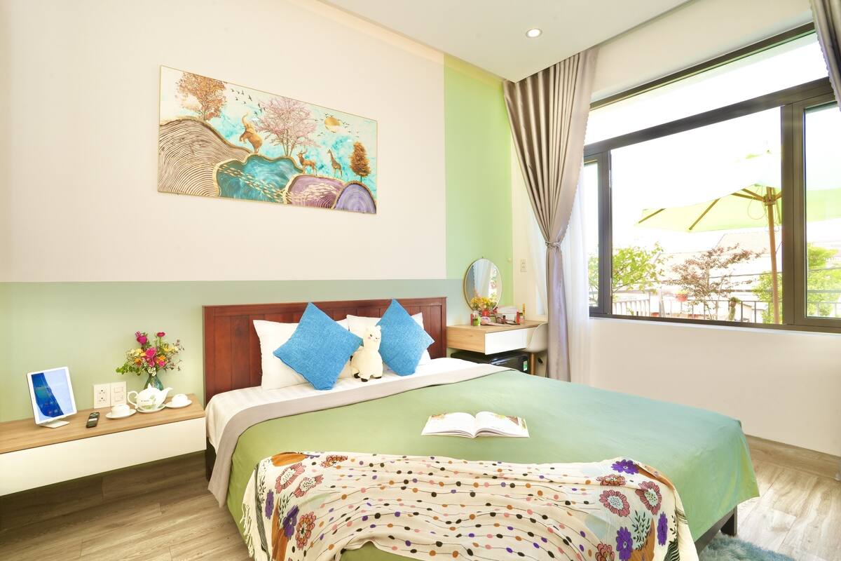 A hotel room with a bed, a bedside table with a lamp, a window with curtains, and a painting on the wall.