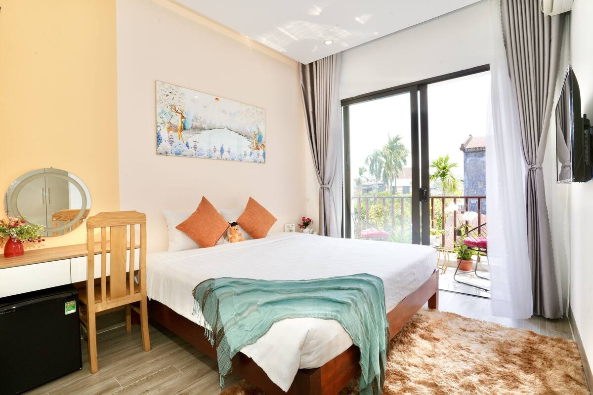 A bedroom with a bed, a desk, a chair, a small refrigerator, a painting on the wall, and a balcony with plants.