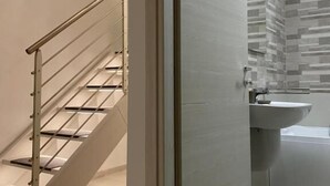A modern bathroom with a glass-enclosed shower, a white sink, and a staircase with metal railings. - Monopoly Sky House (Monopoli)