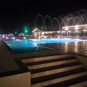 Une piscine nocturne avec des fontaines, des marches menant à la piscine et un bâtiment illuminé en arrière-plan.