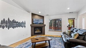 A living room with a fireplace, a large window, a black leather sofa, and a wooden coffee table. - Under the Pines | Quiet Backyard & Neighborhood (Flagstaff)