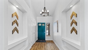 A hallway with white walls, wooden flooring, a teal door, and decorative wooden arrow signs. - Under the Pines | Quiet Backyard & Neighborhood (Flagstaff)