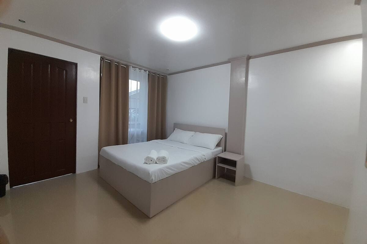 A bedroom with a bed, a nightstand, a door, and a window with curtains.