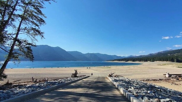Un sendero pavimentado que lleva a una playa, con vista despejada a un lago y montañas al fondo.