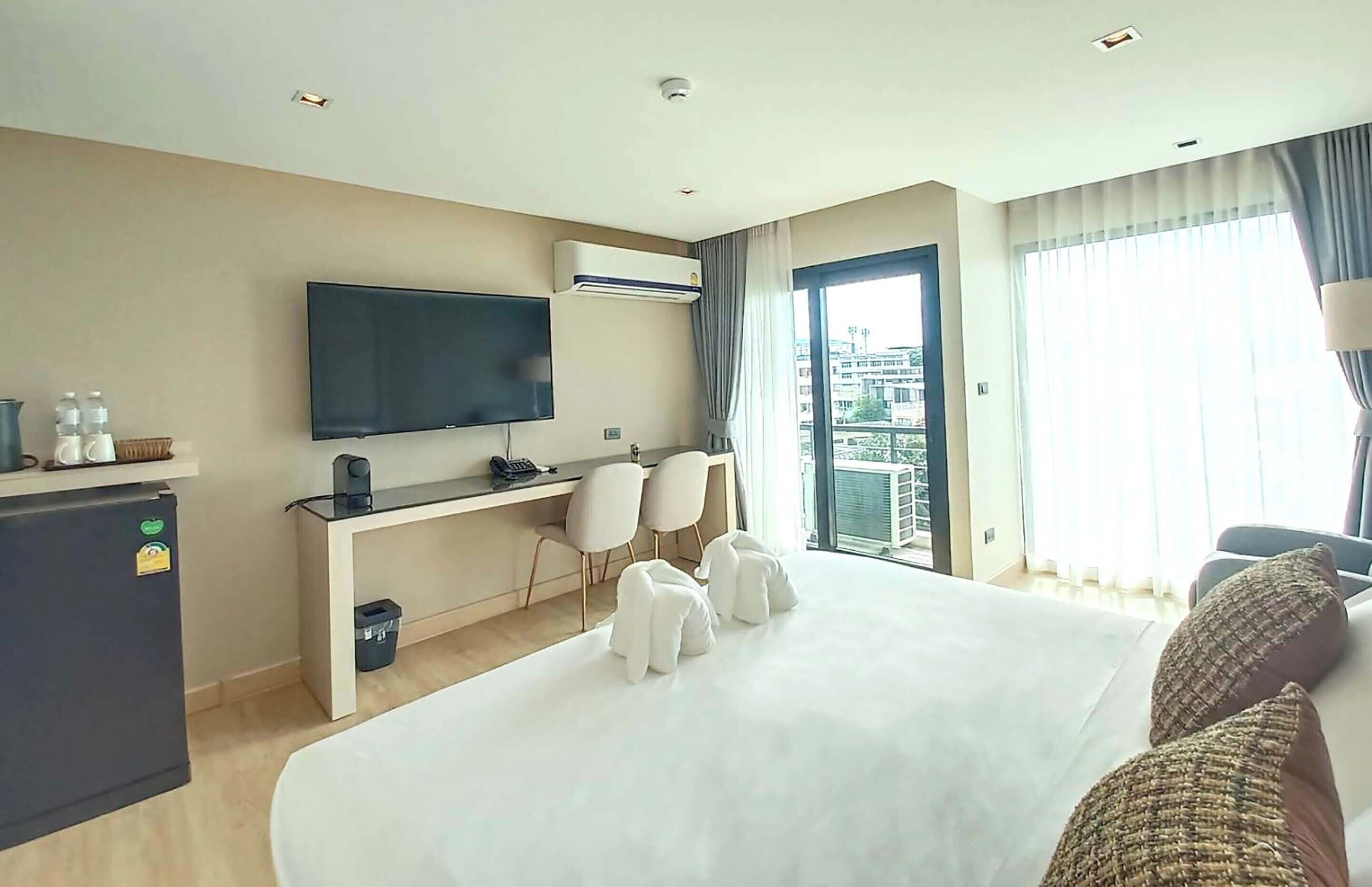 Een hotelkamer met een bed, een bureau met een flatscreen televisie, een koelkast, een prullenbak en een raam met gordijnen.