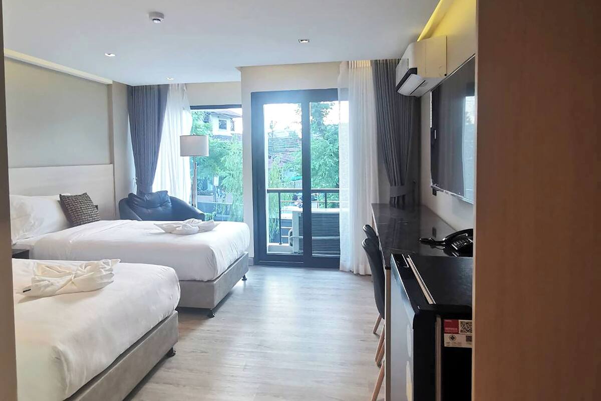 A hotel room with two beds, a desk, a TV, and a balcony with a view of greenery.