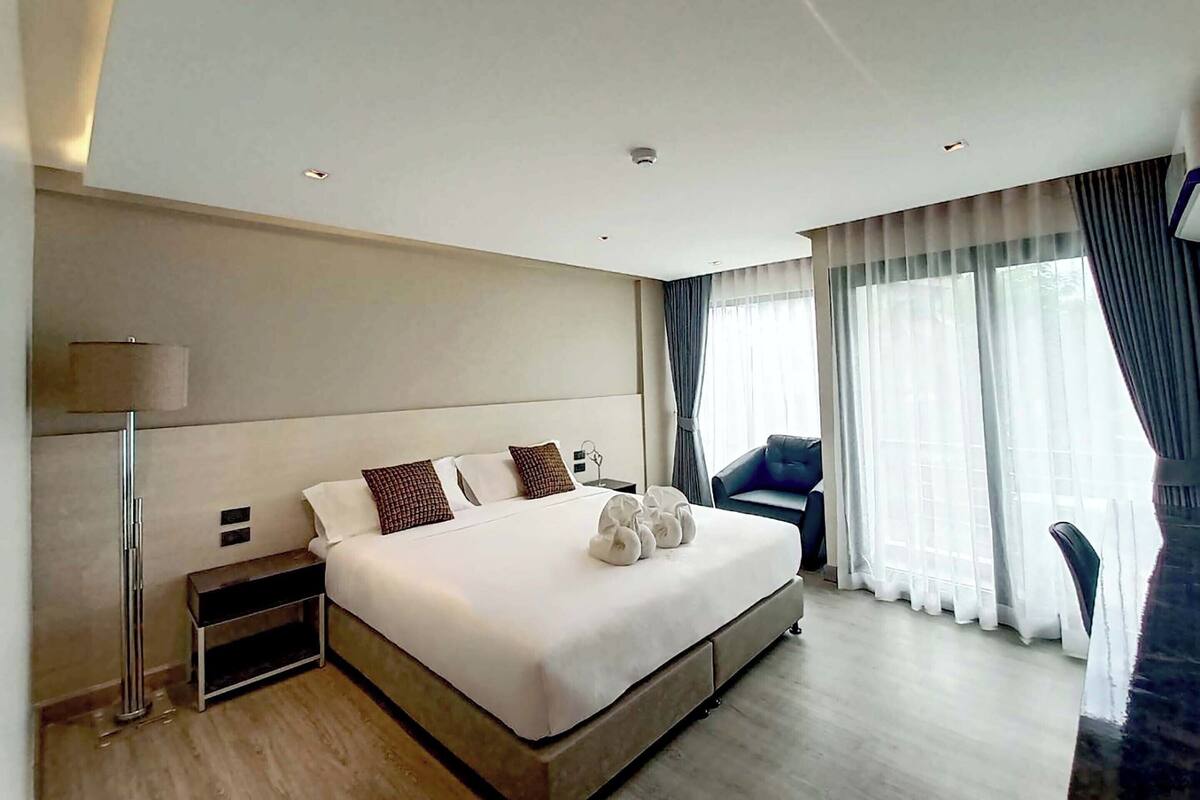 A modern hotel room with a large bed, bedside tables, a lamp, and a view of the outdoors through large windows.