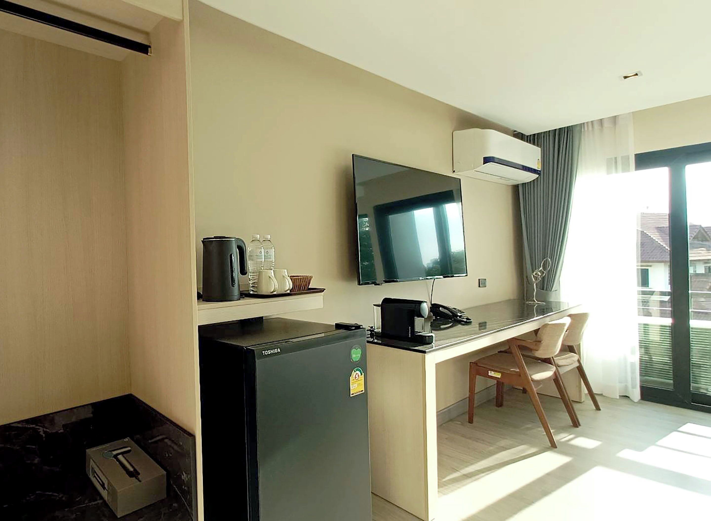 A compact hotel room with a kitchenette, a flat-screen TV, a desk with a chair, and a large window offering a view of buildings.