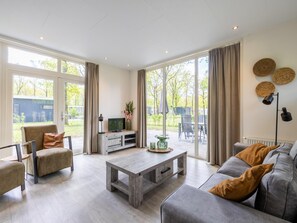 A spacious living room with a grey sofa, a wooden coffee table, and a TV on a wooden stand. Large windows offer a view of the outdoors. - Comfortable chalet in green surroundings (Zorgvlied)