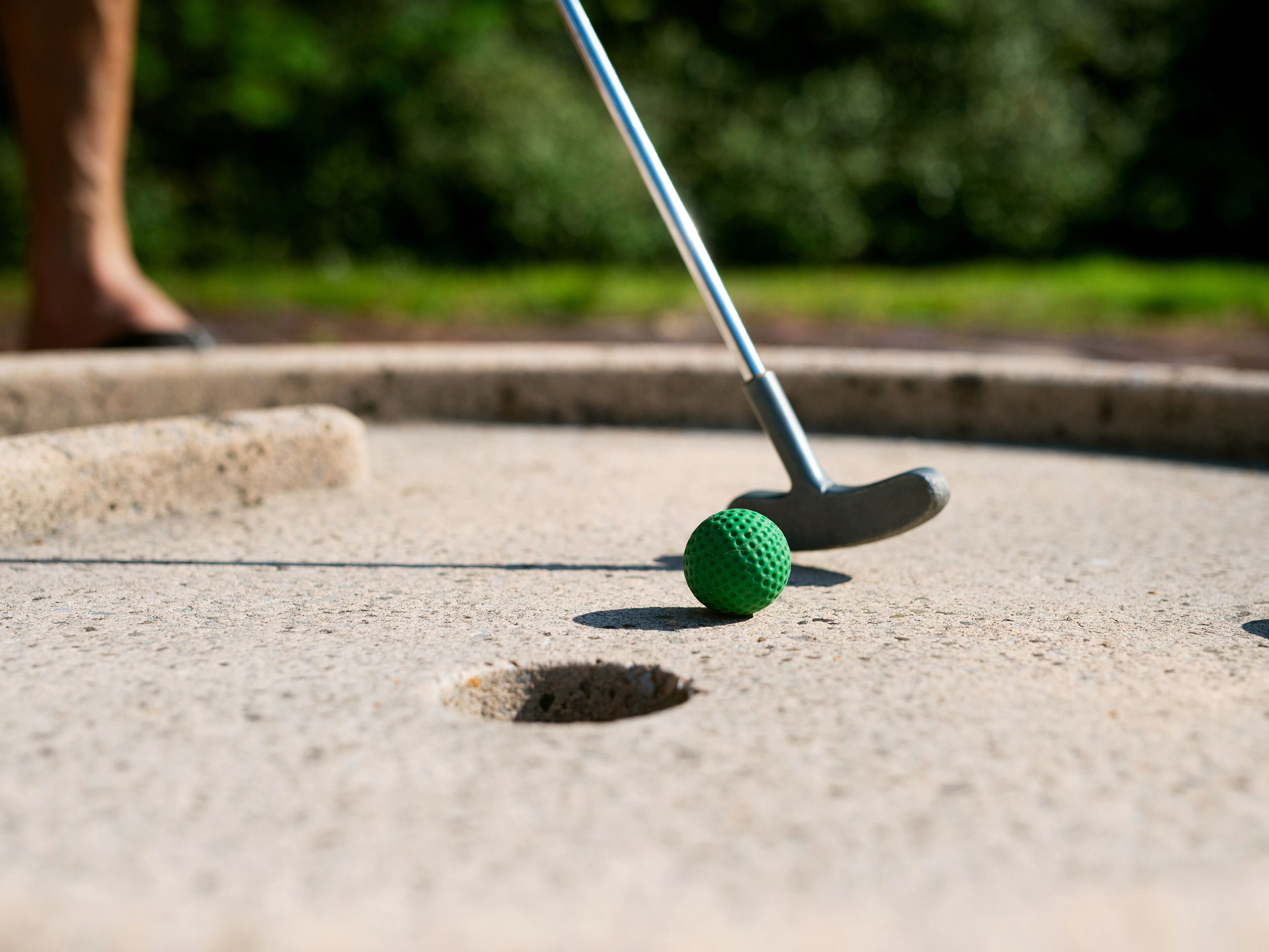 A close-up of a golf ball on a concrete surface with a golf club positioned behind it.