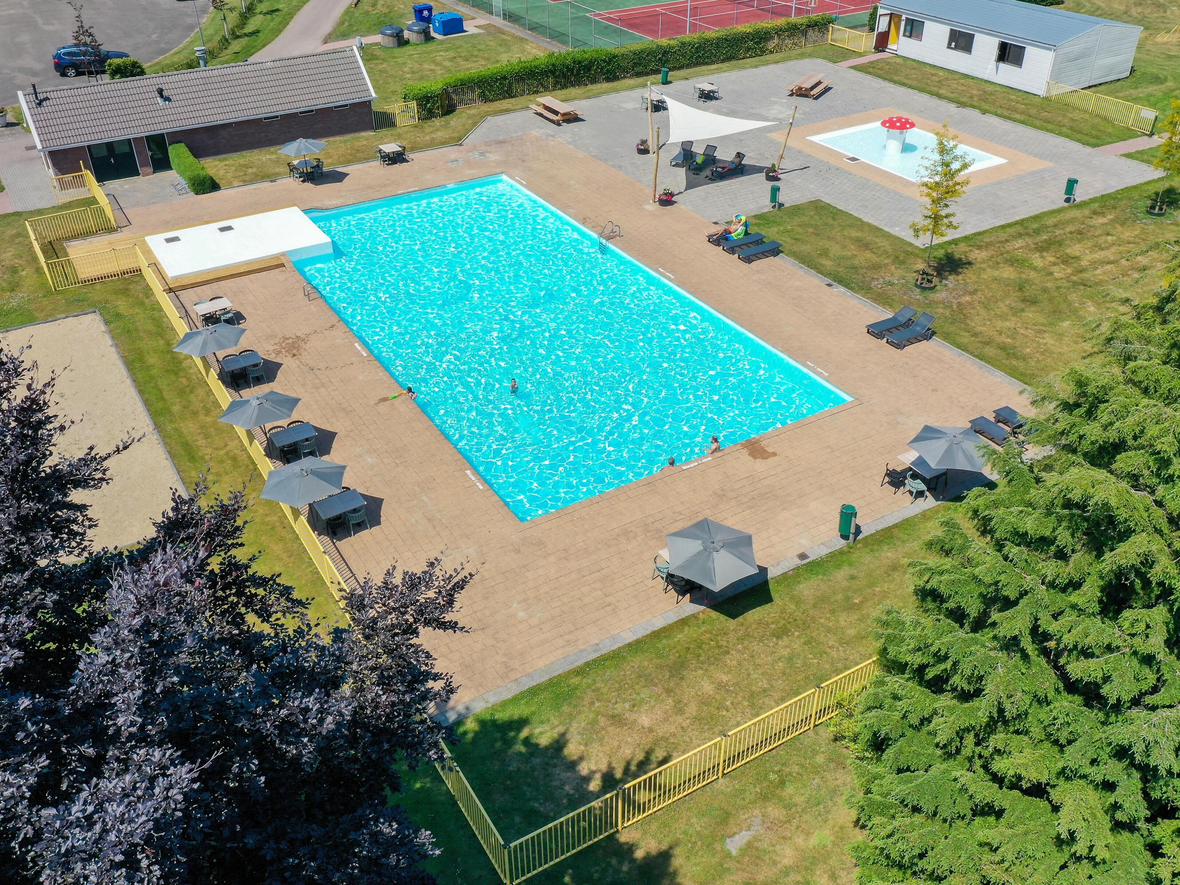 A rectangular swimming pool surrounded by lounge chairs and umbrellas, with a grassy area and a building in the background.