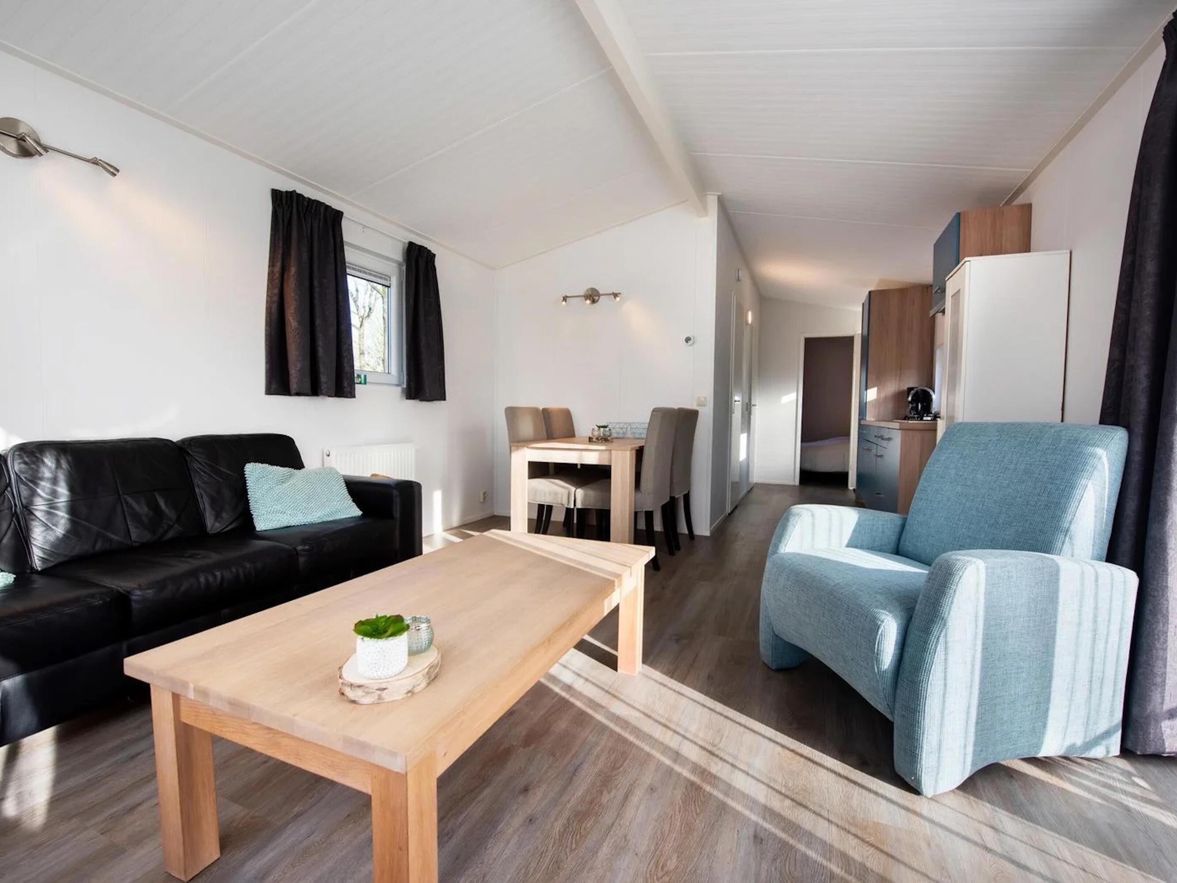 A modern living room with a black leather sofa, a light blue armchair, and a wooden coffee table. There is a dining area with a table and chairs, and a kitchenette in the background.