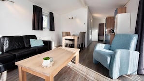 En moderne stue med en sort lædersofa, en lyseblå lænestol og et træbord. Der er et spiseområde med bord og stole, og et tekøkken i baggrunden.