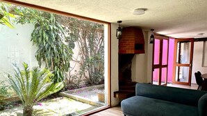 Un salon avec un canapé vert, un plancher en bois, une porte vitrée donnant sur un jardin et une cheminée en briques.