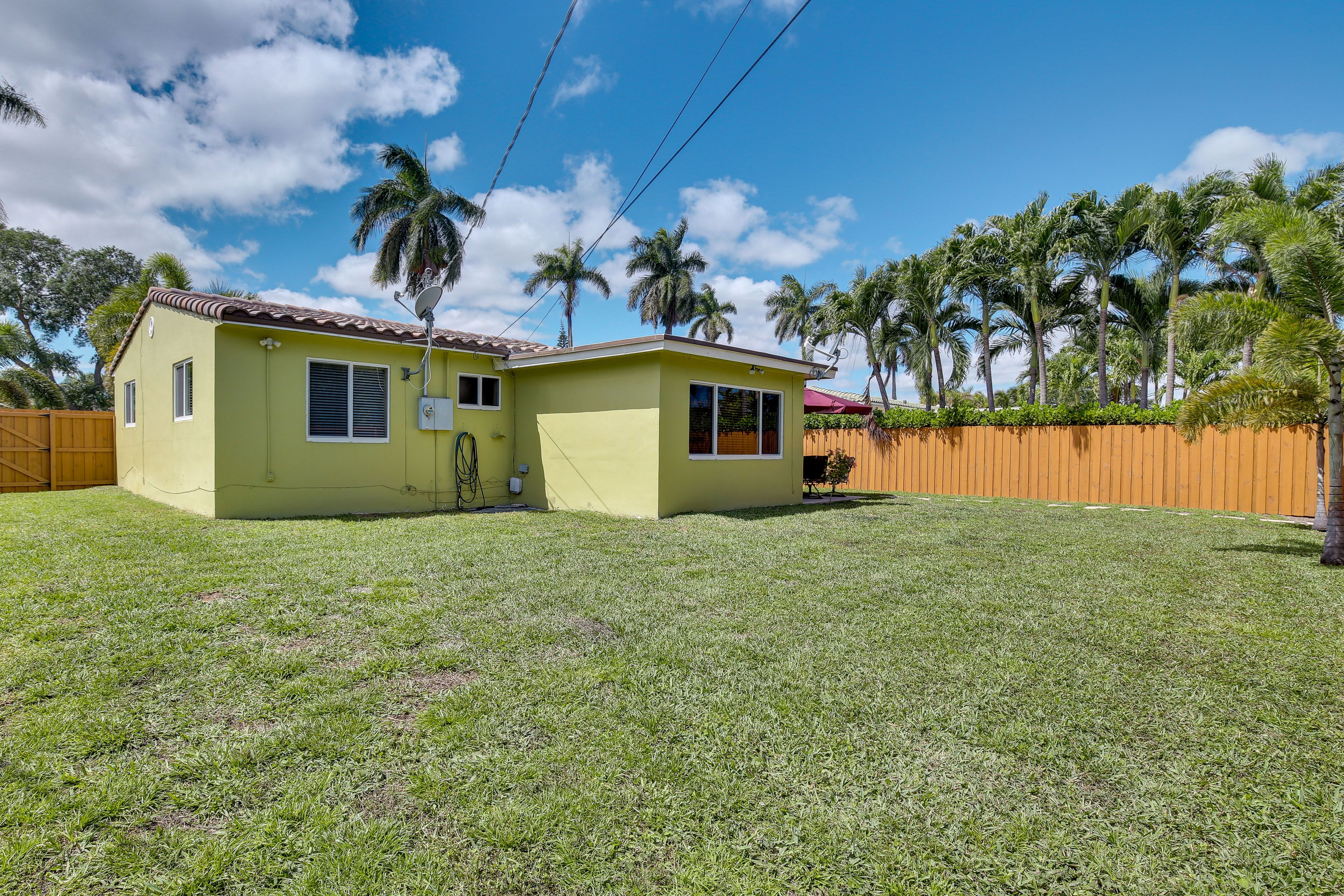 A single-story yellow house with a fenced backyard, surrounded by palm trees.