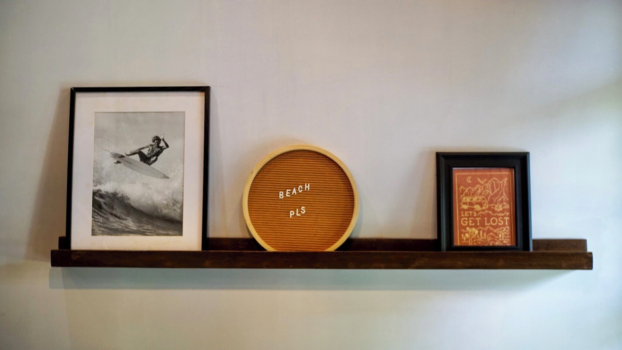 A shelf with three items: a framed photograph of a surfer, a circular wooden board with 'BEACH PL' text, and a framed picture with a mountain scene.