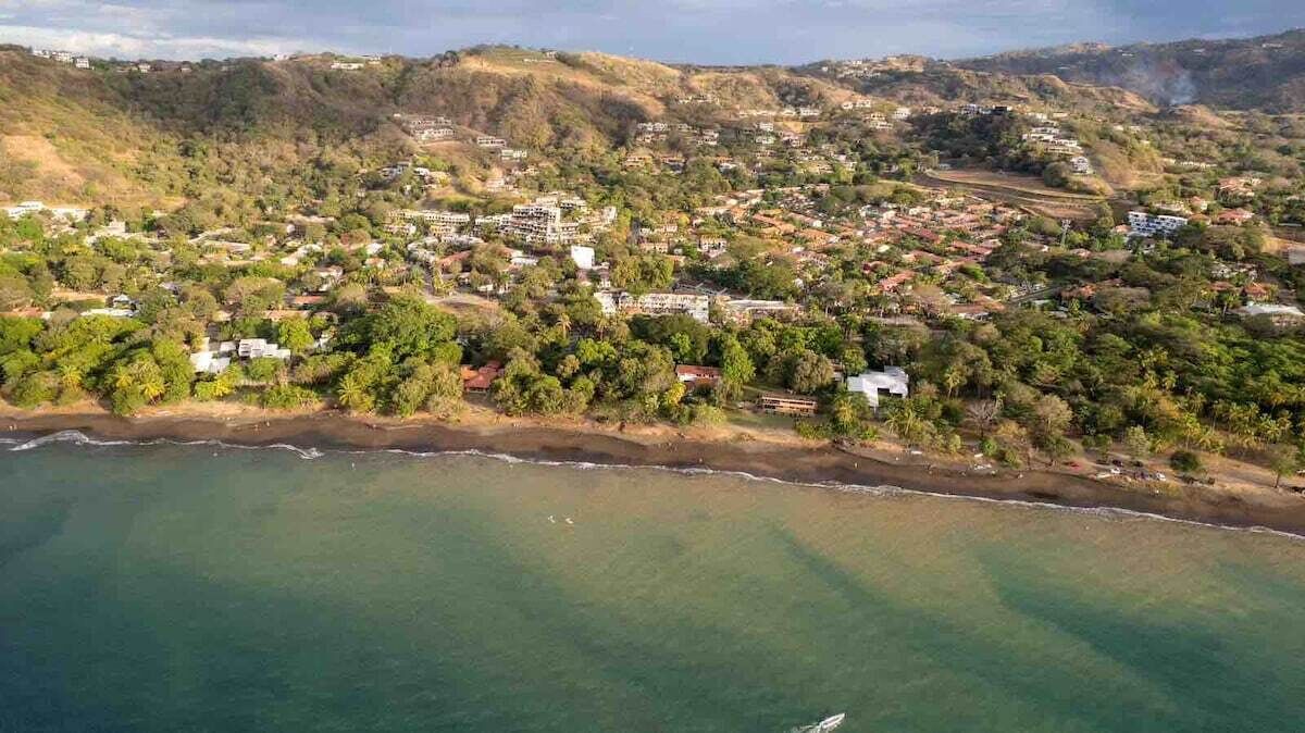 Vista aérea de un pueblo costero con edificios residenciales, una playa y colinas al fondo.
