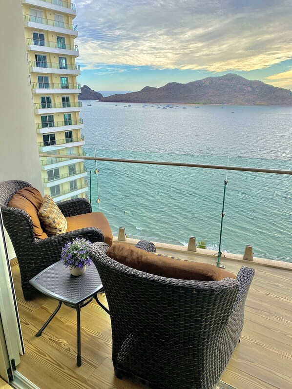 Un balcon agrémenté de meubles en osier, d’une petite table et offrant une vue sur la mer, avec des bateaux et des montagnes en arrière-plan.