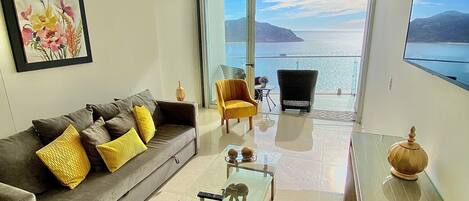 Una sala de estar moderna con sofá, mesa de centro y sillones, un ventanal con vistas al mar y un cuadro en la pared.