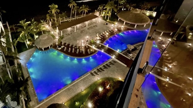 A nighttime view of a resort pool area with a large pool, lounge chairs, palm trees, and a view of the ocean. - Camino al Mar, Apartment on the Beach with Fabulous Amenities Zona Dorada Pure Luxury (Mazatlán)