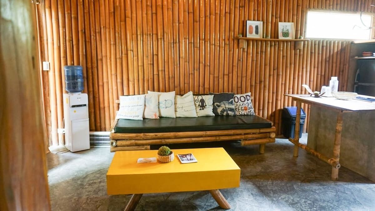 A room with a bamboo wall, a bed with pillows, a small table with a plant, and a water dispenser.