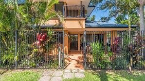 Une maison à deux étages dotée d’un balcon, entourée d’une végétation tropicale luxuriante et d’une clôture en métal.