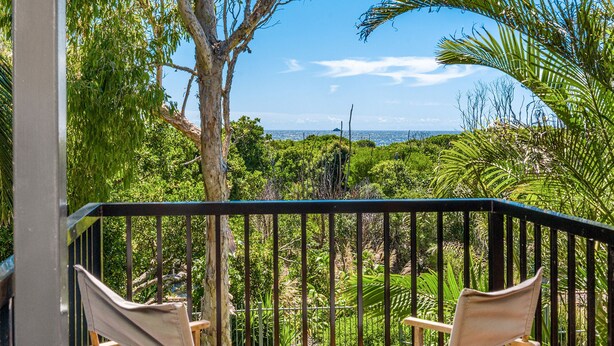 Un balcon avec deux chaises en bois, une rambarde en métal et une vue sur un paysage verdoyant, avec des arbres et un ciel dégagé.