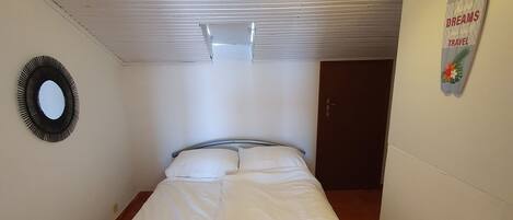 Una cama individual con estructura metálica, ropa de cama blanca, un espejo redondo en la pared y una lámpara en el techo.