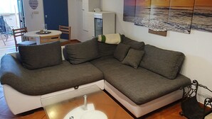 Uma sala de estar moderna com um sofá seccional, uma área de jantar com mesa e cadeiras, e uma pintura grande de um pôr do sol sobre o oceano.