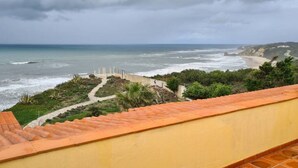 Vista costeira de uma varanda com telhado de azulejos, com vista para o mar e um caminho que leva a uma praia.