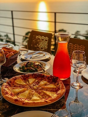 A table set for a meal with a pizza, salad, and a pitcher of red drink, with a sunset view in the background. - Hotel Hills Kvariati (Batumi)