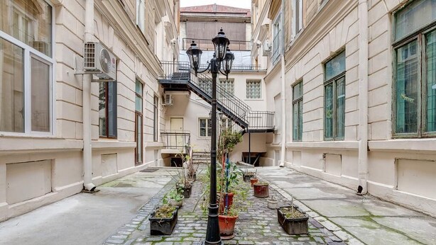 A narrow cobblestone courtyard with a central staircase, surrounded by buildings with large windows and potted plants.