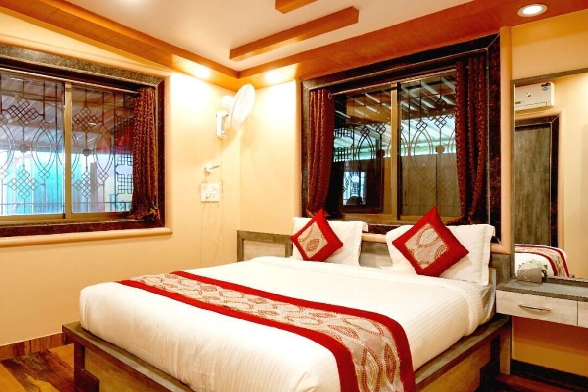 Quarto de hotel com cama, mesa de cabeceira, espelho, janela com cortinas e luminária na parede.