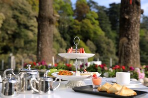 Un soporte para postres de tres niveles con fresas y un vaso de jugo sobre una mesa al aire libre, con árboles y flores al fondo.