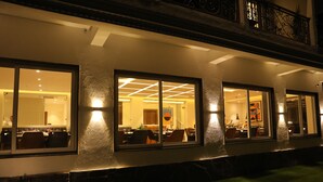 L’extérieur d’un hôtel de nuit, avec des fenêtres illuminées, une façade en pierre et un balcon orné de balustrades en fer forgé.