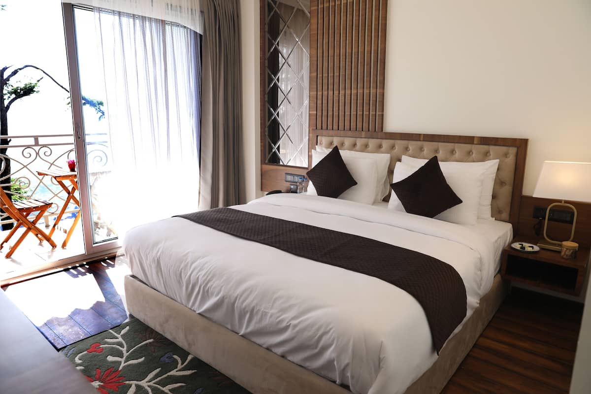 Habitación de hotel con una cama grande, un cabecero de madera, una mesita de noche con una lámpara y vistas al exterior a través de una puerta corrediza.